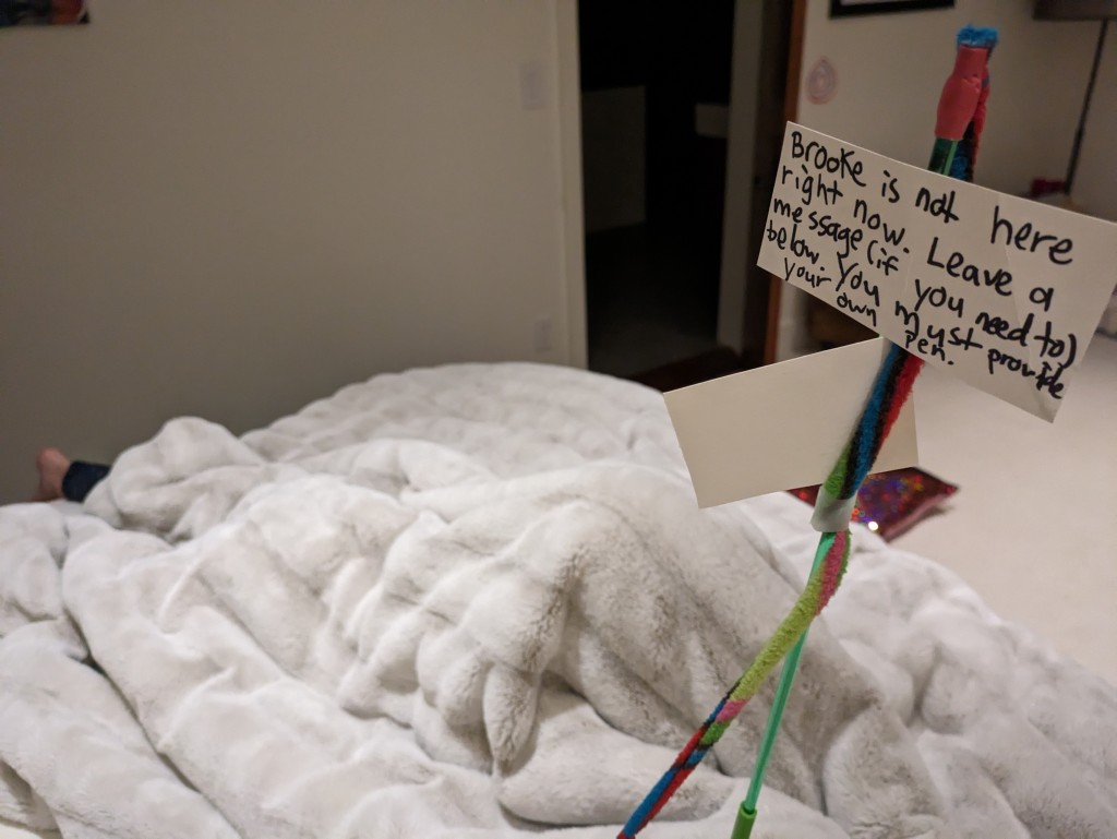 Sign on bed that says "Brooke is not here right now. Leave a message (if you need to) below. You must provide your own pen." A foot is poking out from under the covers.